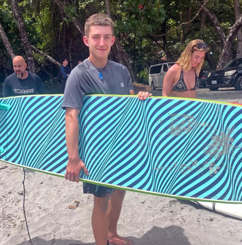 A young man stands on a sandy beach, holding a blue and black striped surfboard. He is positioned in front of a woman in a bikini top and a man in a dark t-shirt. In the background, there are trees, a white van, and a black SUV. The scene suggests a beach outing, possibly related to surfing or water sports.